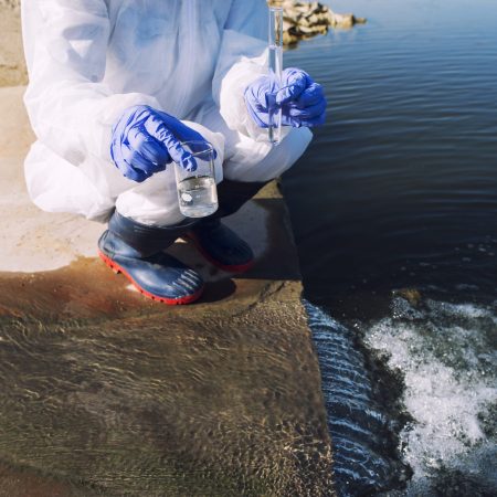 Unrecognizable ecologist standing where sewage waste water meets the river and taking samples to determine level of contamination and pollution.