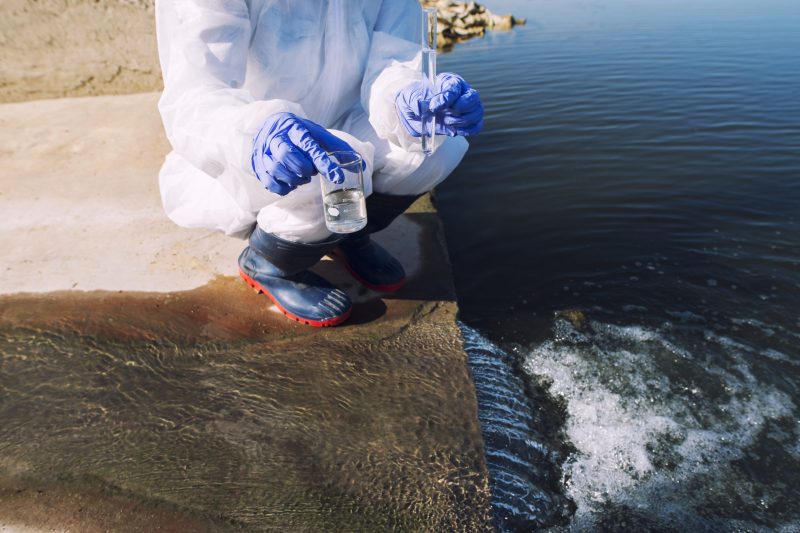 Unrecognizable ecologist standing where sewage waste water meets the river and taking samples to determine level of contamination and pollution.